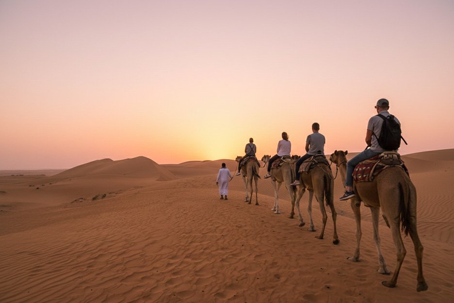 Una carovana di cammelli attraversa le dune di sabbia dorata del deserto di Wahiba Sands durante un suggestivo tramonto, Oman
