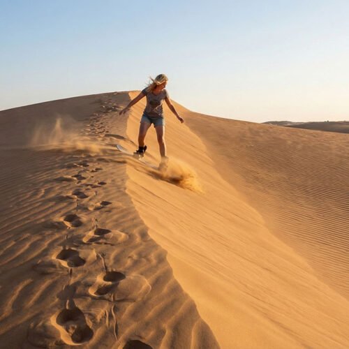 Un viaggiatore che fa sandboarding scivolando lungo le alte dune di sabbia dorata del deserto di Wahiba Sands, Oman